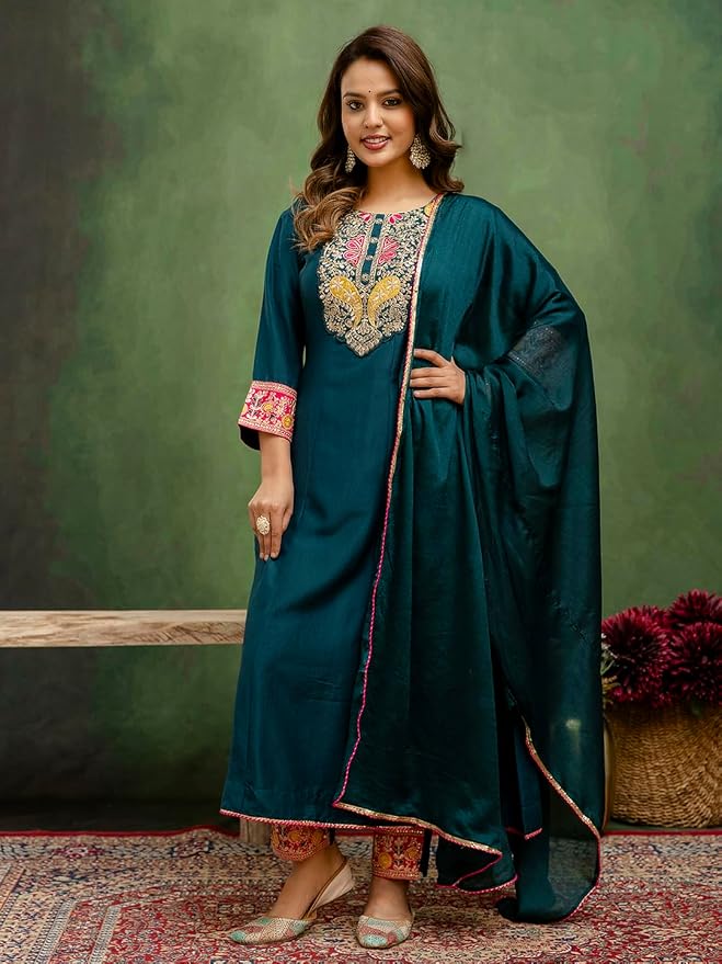 A woman models a dark teal green ethnic kurta set with intricate gold, pink, and green embroidery on the neckline and cuffs, paired with a matching sheer dupatta, set against a textured green studio backdrop.